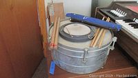 Two metal timbales stacked with drumsticks and a blue Suzuki recorder case on top, sitting next to a keyboard.