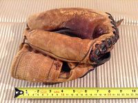 Top side of brown leather vintage catchers glove with partially visible faint signature and black lacing, placed beside yellow measuring tape indicating length approximately 11-12 inches.