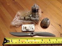 Overview photo of disassembled OS Max 3 OS 15 model airplane engine parts on a wooden surface including engine block, propeller, spinner, and a switch plate with ruler for size reference.