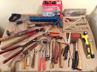 Photo showing the lot of mixed tools including the 3M window insulator kit, grease gun, axes, pliers, screwdrivers, saws, and other hand tools spread out on a cardboard surface.