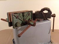 Overall view of both bench vices on display, showing the Rae Canada vice with painted body and wooden jaws, and the Hercules vice with rusted steel body.