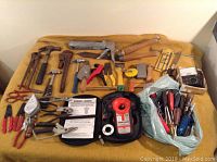 Overview of hand tools spread out on a yellow cloth, includes hammers, wrenches, pliers, screwdrivers, scissors, a meter and manual, and a caulking gun.