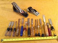 Photo showing two metal wood planes and fourteen wood chisels with a tape measure indicating length.