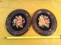 Pair of oval antique wood picture frames on yellow cloth with measuring tape showing about 15 inch width. Both frames contain floral needlepoint art on black fabric.