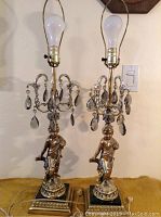 Pair of 1960s cherub table lamps with hanging prisms and brass fittings, bulbs installed, shown from front angle on surface.