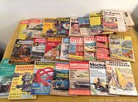 Wide shot of approximately 30 vintage magazines spread out showing various titles including Mechanix Illustrated, Popular Science, and others with colorful mid-century themed covers featuring cars, engines, and tools.