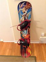 Full view of snowboard standing vertically against wall showing length and attached bindings.