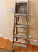 Photo of 5 foot aluminum step ladder showing 4 steps and top platform, against a beige wall on wood flooring.
