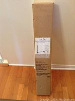 Full box standing upright against a wall on wood floor. Box label shows the product image and dimensions.