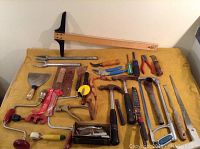 Full lot of vintage hand tools laid out on yellow fabric showing various items described including wooden T-square, smelting ladle, art knife, hammers, saws, pliers, tape measure.