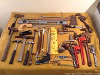 Full overhead view of assorted hand tools arranged on yellow cloth including hammers, saws, pipe wrenches, levels, axes, and hand drills.