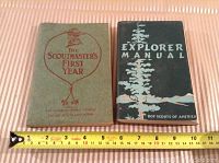 Two vintage Boy Scout books lying flat on a striped surface with a measuring tape showing dimensions. Left book is titled 'The Scoutmaster's First Year', right book titled 'Explorer Manual'.