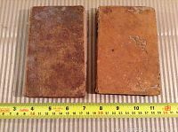 Two old hardcover books side by side on a cloth surface with a measuring tape showing sizes. The left book has a dark brown cover and the right book a lighter tan cover, both with visible wear and stains.
