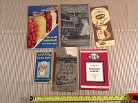 Seven vintage cook and canning books arranged on a cloth surface, includes nursery blue Magic Cook Book, How to Can Finer Fruits, Canning at Home, rheumatic ailments guide, and more