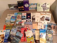 Wide shot showing over 30 travel brochures and maps spread on a table with various Canadian and international locations depicted and vintage 1960s-70s design.