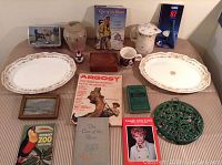 Lot items arranged on a table including books, two oval platters, mugs, printed materials, small framed pictures, and trivet.