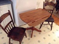 Photo showing the wooden table with one drop leaf partially down and two matching wooden chairs placed on either side on a light patterned floor.