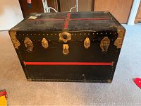 Front view of black vintage trunk with red bindings and brass corner accents, showing rivets and metal latches.