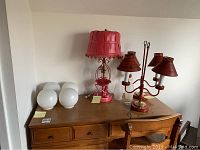 Image showing two red tole metal lamps with stenciled shades, pink basket lamp, and multiple glass lamp shades on wooden table.