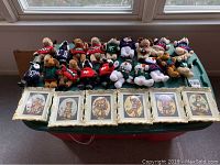 Photo of all 19 stuffed animals in colorful Christmas sweaters arranged in rows behind 6 framed Hummel prints on a table under a window.