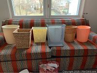 Six assorted waste baskets displayed on a patterned textile sofa. Various materials including plastic, woven, and wire mesh visible.