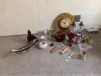 Photo showing variety of decorative items including brass candle holders, crystal vases, wooden horse head sculpture, religious statues, wall clock, bowls, and wristwatch arranged on carpet floor.
