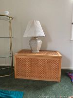 Whole view of woven reed and wood chest with ceramic floral lamp and white pleated shade on top