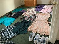 Photo showing multiple women's dresses and skirt outfits laid out on green carpet, along with two pairs of white sneakers and a pink satin clutch with pumps.