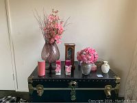 Full view of the lot arranged on dark green trunk showing pink faux flowers in purple vase, pillar candles, small faux flower arrangement in vase, wooden jewelry case, decorative candle holders, and round basket