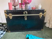 Front view of black vintage trunk with brass latches, hinges, and leather handles on carpeted floor.