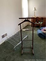 Wooden suit caddy standing with metal shoe racks on carpeted floor near a chair and lamp.