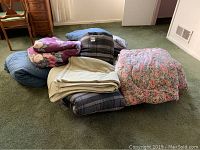 Photo showing multiple bedding items including comforters with floral and plaid patterns, a beige blanket, and blue bedding items.