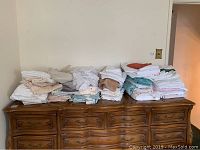 Stacks of assorted bed linens, towels, and linens folded on a wooden dresser.