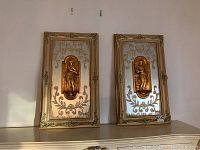Pair of rectangular wall panels with gold relief Grecian maidens and ornate frames displayed against a wall.