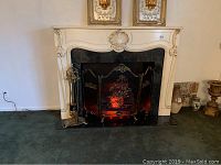 Full view of cream-colored ornate fireplace surround with black insert showing glowing faux logs, and decorative metal fireplace screen in front.