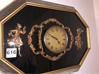 Front view of octagonal wall clock with gold Rococo style figures and scrolls.