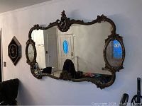 Full frontal image of the large ornate wall mirror with gold tone frame and two oval side mirrors hung on wall.