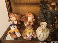 Two fall-themed girl figurines with pumpkin hats and brooms, two Easter bunny plush figurines, and a small white vase with gold bow on wood shelf