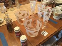 Photo showing four etched highball glasses with frosted leaf designs and two leaf-decorated bowls with gold/brown detailing, arranged with multiple other glass and ceramic items on a wooden table.