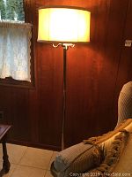 Four bulb brass standing lamp near a lace-curtained window, lamp lit showing warm light through the fabric shade.