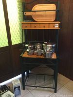 Full view of bakers rack with metal frame, wooden shelves and three small drawers. Includes several stainless steel pots and pans, not included in lot.