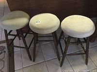 Three wooden stools with round light-colored upholstered cushions placed side by side showing worn wood legs and frame.