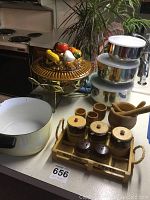 Photo showing ceramic tureen with vegetable-themed lid on brass stand, white enamel crock, wooden pedestal cups, wooden mortar and pestle, condiment tray with six labeled jars with wooden lids on handle carrier.
Also shows three stainless steel mixing bowls with white plastic lids stacked.