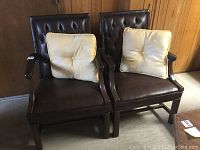 Pair of brown vinyl executive office armchairs together with ivory cushions on seats, brown wood panel wall background.