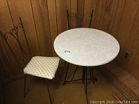 Round cafe table with white marbled top and black metal legs alongside one metal chair with decorative back and worn cushioned seat.