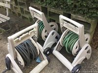 Three white wheeled hose reels with hoses coiled on them, positioned outdoors on stone/brick ground with a concrete wall background.