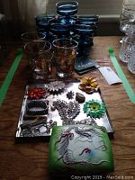 Multiple glassware items and jewellery displayed on a table, including blue footed fruit nappies, tumblers with gold designs, vanity box, and various brooches.