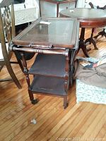 Side view of tea wagon showing three wooden shelves, turned legs, and large spoked wheels at rear with smaller caster wheels at front on hardwood floor.