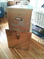 Two metal storage boxes stacked on wooden floor; medium box on top with handle and lock, larger box below with clamps.