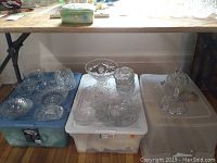 Large assortment of cut glass trays, bowls, butter dishes, and fruit nappies placed on plastic bins under a wooden table.